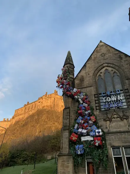 A tower of wrapped presents climbing a stone church, with Edinburgh Castle behind